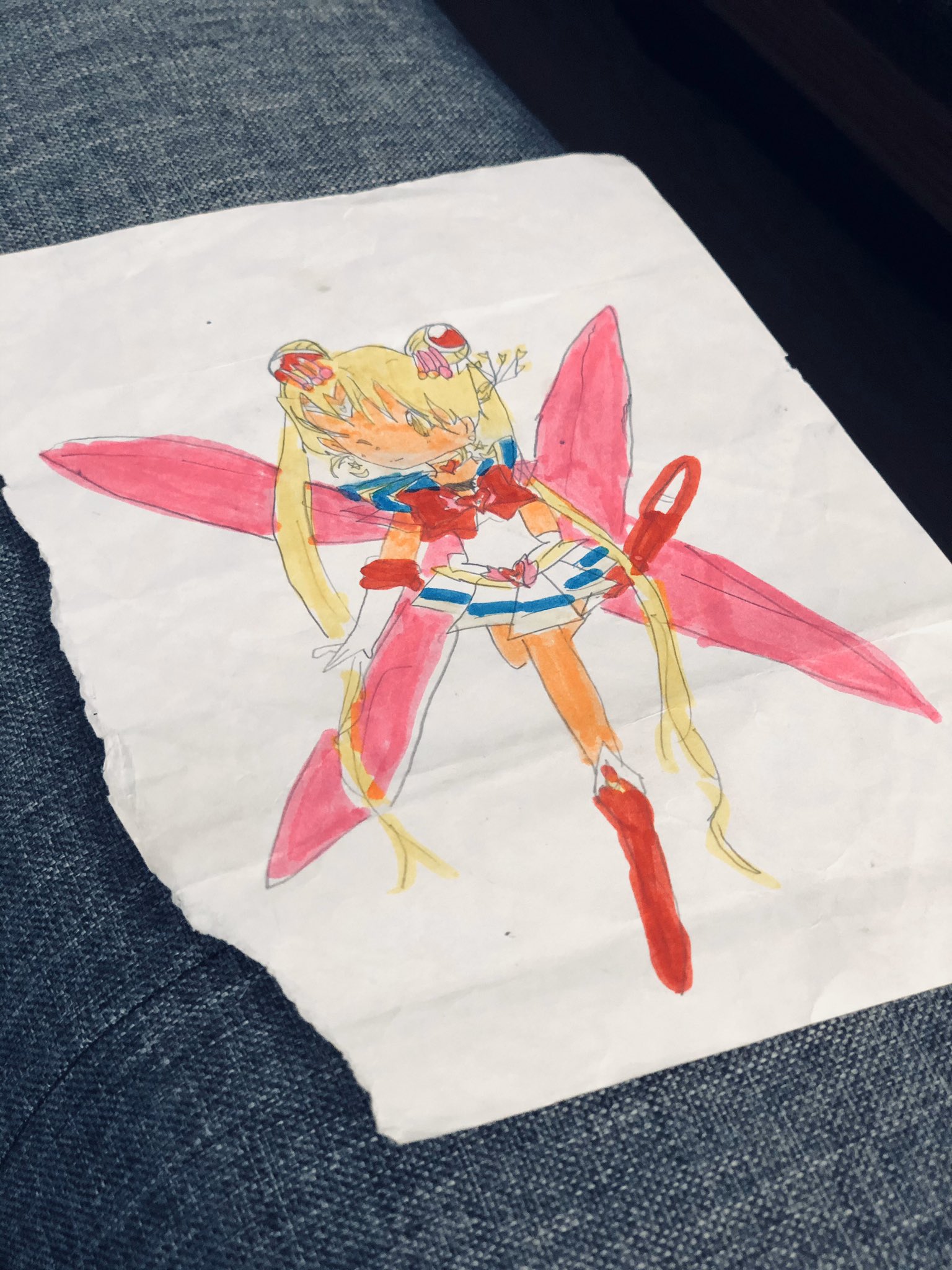 a child's drawing of sailor moon in marker and colored pencil on a piece of white paper. the drawing is of the character in her transformed state, with her pink and white costume, blonde hair in odango (buns) with red hair ties, and large pink wings. she is positioned slightly turned with her left leg extended and holding her right hand up in her signature pose.  the drawing is done on a piece of white paper with a torn corner, laying on a dark blue jean background.