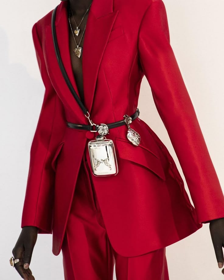 a woman wearing a red suit with a black belt and jewelry. she is wearing a necklace and a ring. she is also carrying a purse.