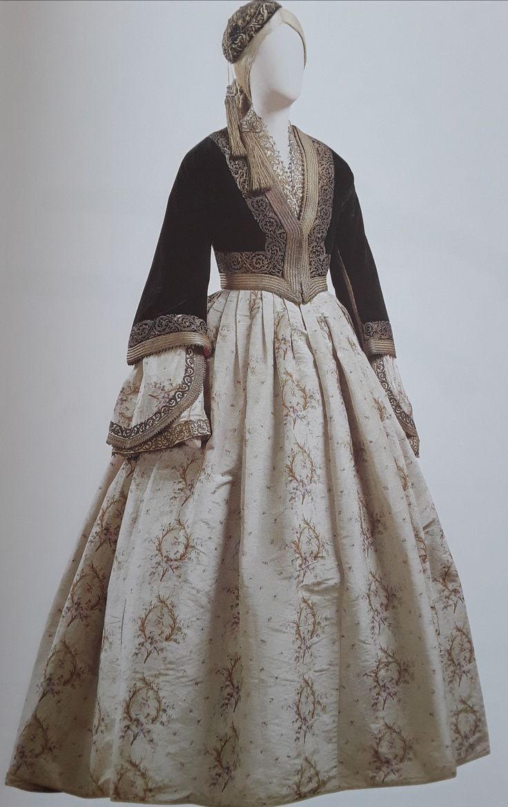 a fulllength studio shot of a mannequin wearing a traditional garment. the garment consists of a dark, possibly black velvet jacket or bodice with gold embroidery along the edges and sleeves, and a full, lightcolored skirt with a floral pattern. the mannequin also wears a matching headpiece, possibly a bonnet or cap, made of the same fabric and embroidered with gold. the mannequin's skin tone is white. the background is a plain, light color.