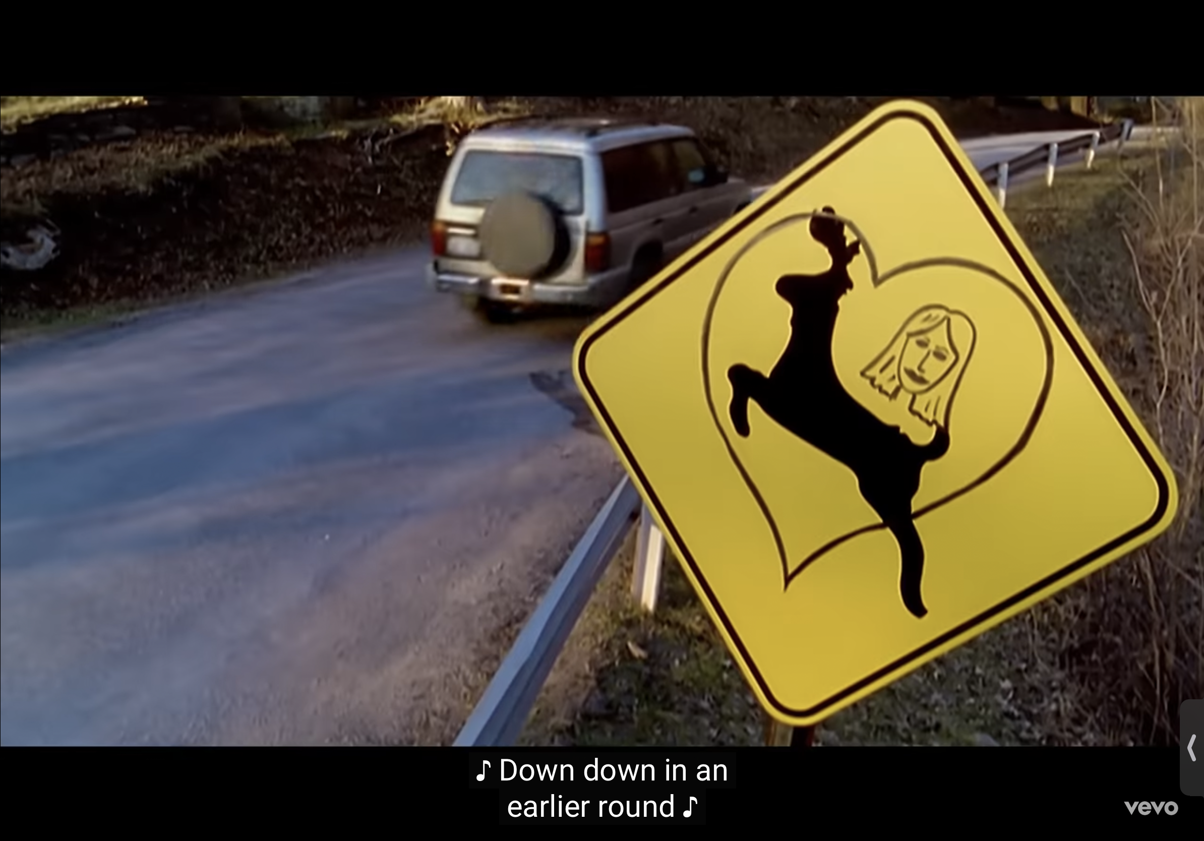 a yellow warning sign with a black silhouette of a deer inside a heart shape is shown in the foreground. the deer has a woman's face. it is on the side of a rural road. a car drives by in the background. the sign is a warning for drivers.
