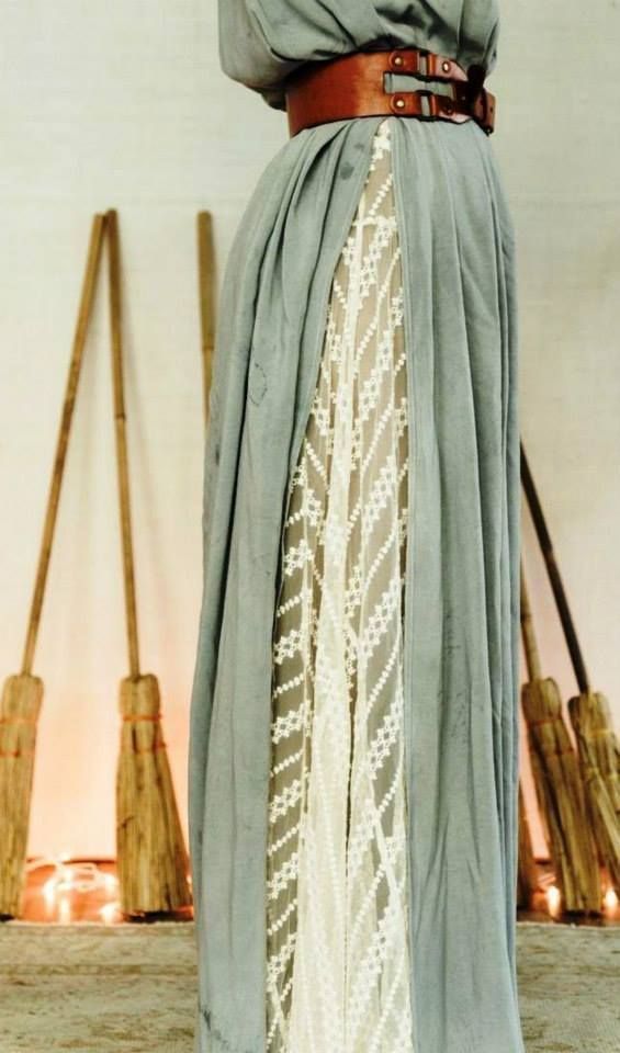a woman is wearing a gray skirt with a brown leather belt. there is a white lace panel running down the center of the skirt. in the background, there are several brooms leaning against a wall, and some candles are lit on the floor.