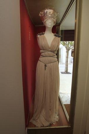 a closeup shot of a mannequin dressed in a long, beige pleated gown with an empire waist.  the gown is adorned with a silver crownlike headpiece and a keyshaped pendant. the mannequin is positioned in a store window display against a red wall. the visible portion of the store interior is subtly included.