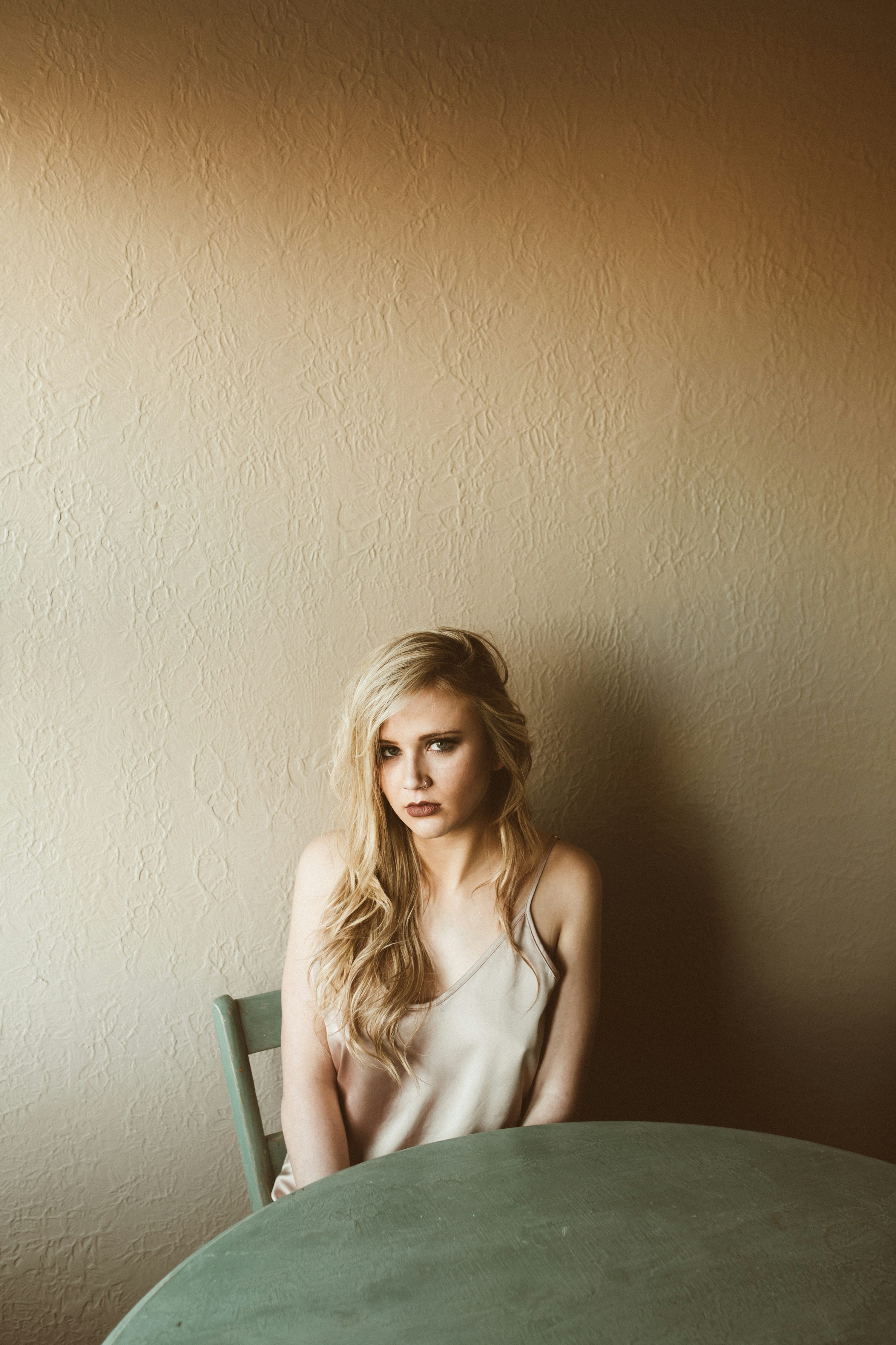 the image is a portrait of a young blonde woman with long hair. she is sitting in a wooden chair at a round table, against a beige textured wall. she is wearing a light beige tank top and appears serious or pensive. the overall mood is calm and quiet. the lighting is soft and natural, creating a minimalist and somewhat melancholic atmosphere. the focus is primarily on the woman, with the background and setting playing a supportive role in conveying the mood.