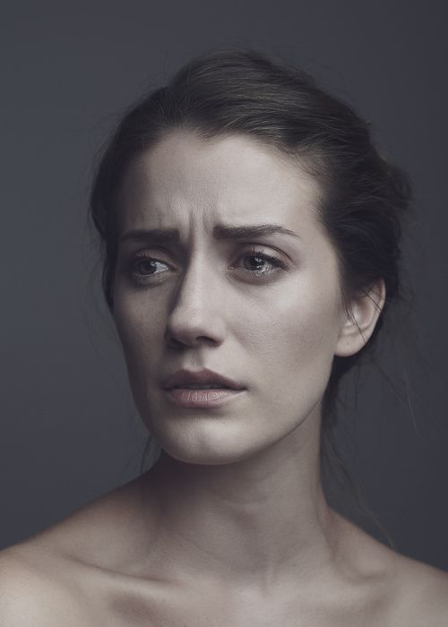 a closeup portrait of a woman with a sad expression. she appears to be on the verge of tears or is actively crying, indicating emotional distress. the image conveys feelings of sadness, vulnerability, and possibly anxiety or worry. the background is a muted grey, which helps to focus attention on the woman's face and emotional state.
