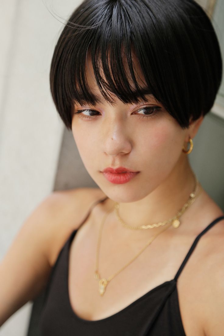 a close up portrait of a young asian woman with short black hair and bangs. she's wearing a black tank top and gold jewelry, including a choker necklace and earrings. the woman is looking directly at the camera with a slightly serious expression.