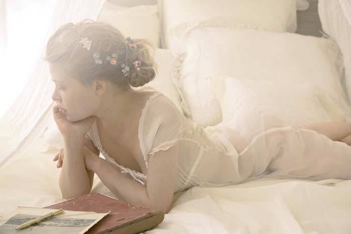a young girl lies on a bed with white linens, appearing thoughtful and perhaps daydreaming. she is wearing a sheer, white dress and has flowers woven into her hair, which is styled in a bun. a book and pen rest on the bed near her, suggesting she may have been reading or writing. the scene is bathed in soft, natural light.