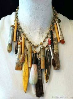 a closeup shot of a unique necklace made with paintbrushes, golden beads, and other trinkets. the necklace is worn by a person. the paintbrushes are of various sizes and colors. the necklace is a creative and stylish accessory.  it’s a boho style necklace.