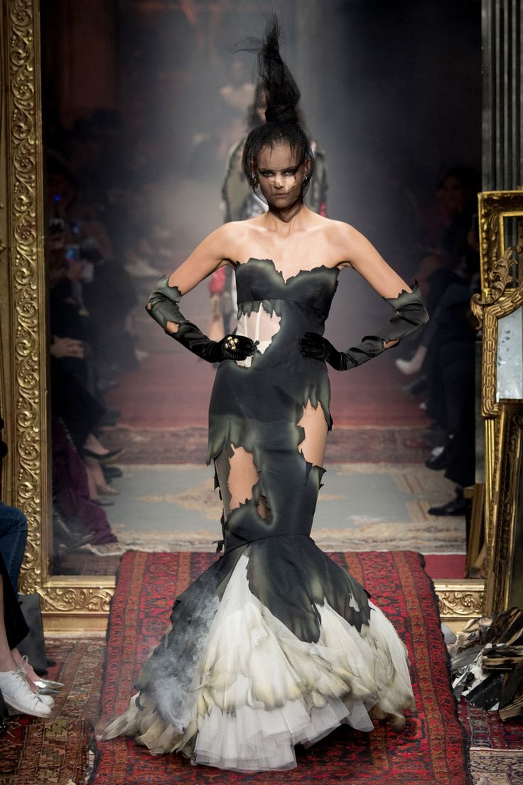 a fashion model is walking the runway in a black and white haute couture gown with a large white fluffy skirt and black sheer top, accessorized with black gloves. the model is wearing her hair up in a bun with long black strands, and is looking straight ahead at the camera as she walks. the runway is red, with ornate gold mirrors framing the sides of the image.