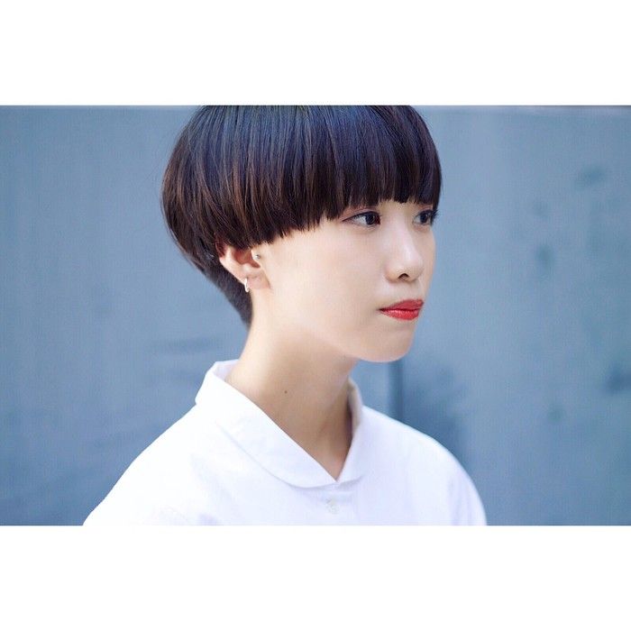 a young woman with short black bob haircut with bangs, wearing a white collar shirt, looking to her right side. she is wearing a small earring on her left ear. her skin is clear and she has bright red lips.  the background is a light blue wall. the picture is taken in a studio setting and is likely taken by a professional photographer.