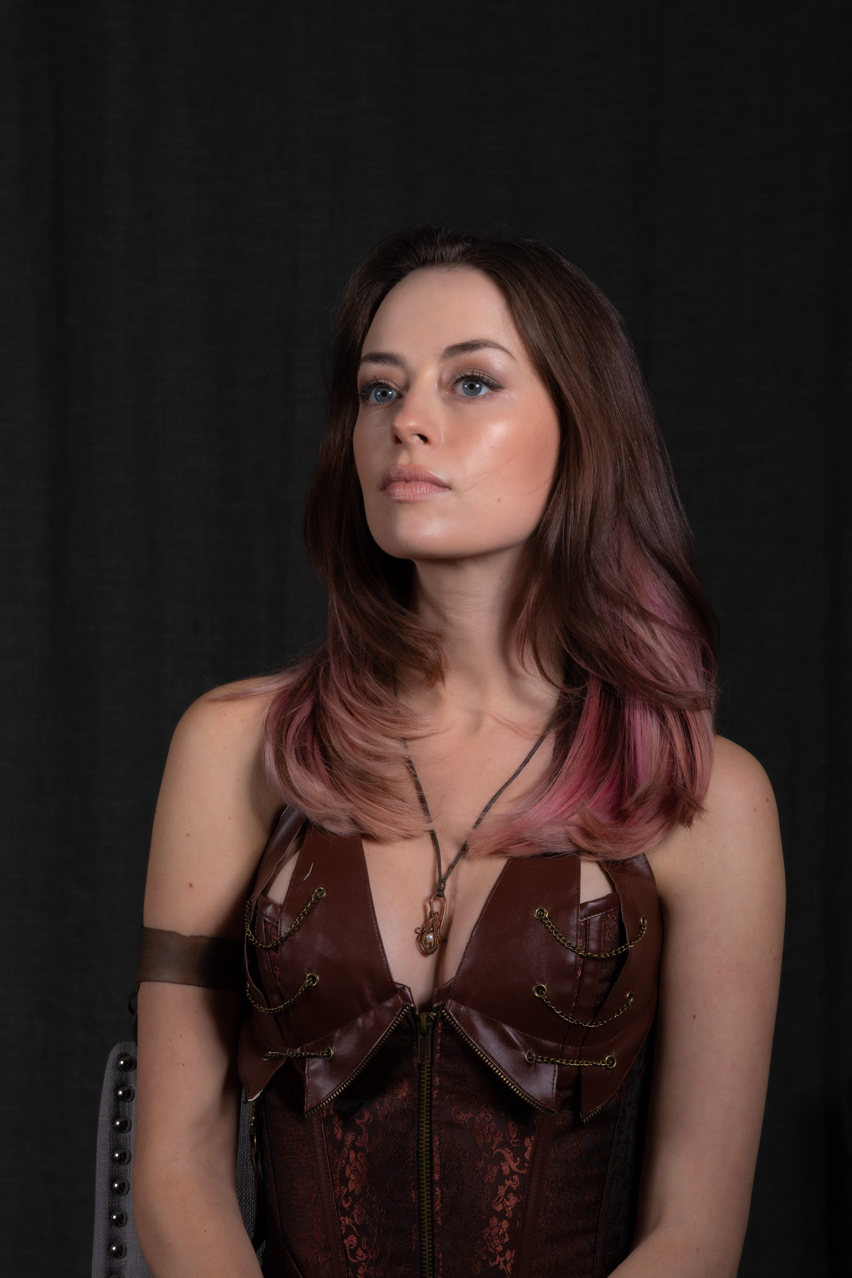 a young woman with brown hair and streaks of pink poses against a black backdrop. she wears a brown steampunkinspired corset with intricate detailing and a matching arm cuff. a delicate necklace with a pendant rests on her chest. the studio lighting casts a soft glow on her face, highlighting her brown eyes and serious expression.  she exudes confidence and a touch of rebelliousness.