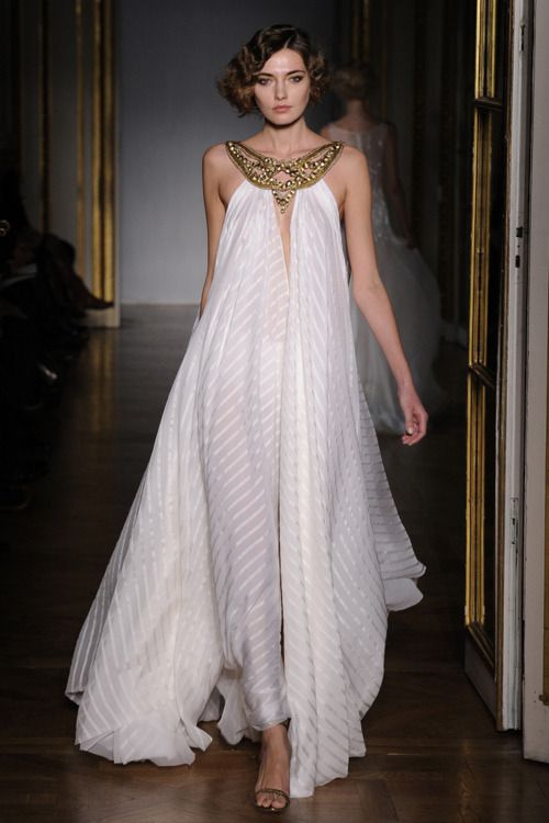 a woman wearing a white gown with gold jewelry walks down a runway. she has a beautiful, elegant, and sophisticated look. the gown is designed with a flowing silhouette, and has a large, gold necklace, which is very stylish. she is wearing high heels, which are also gold. the model's hair is styled in a beautiful and chic manner. the background is a blurred image of the runway and other models. this is a fashion show, and this model is the highlight of the event. the model is looking confidently down the runway and exudes confidence. this is a very beautiful and glamorous image.