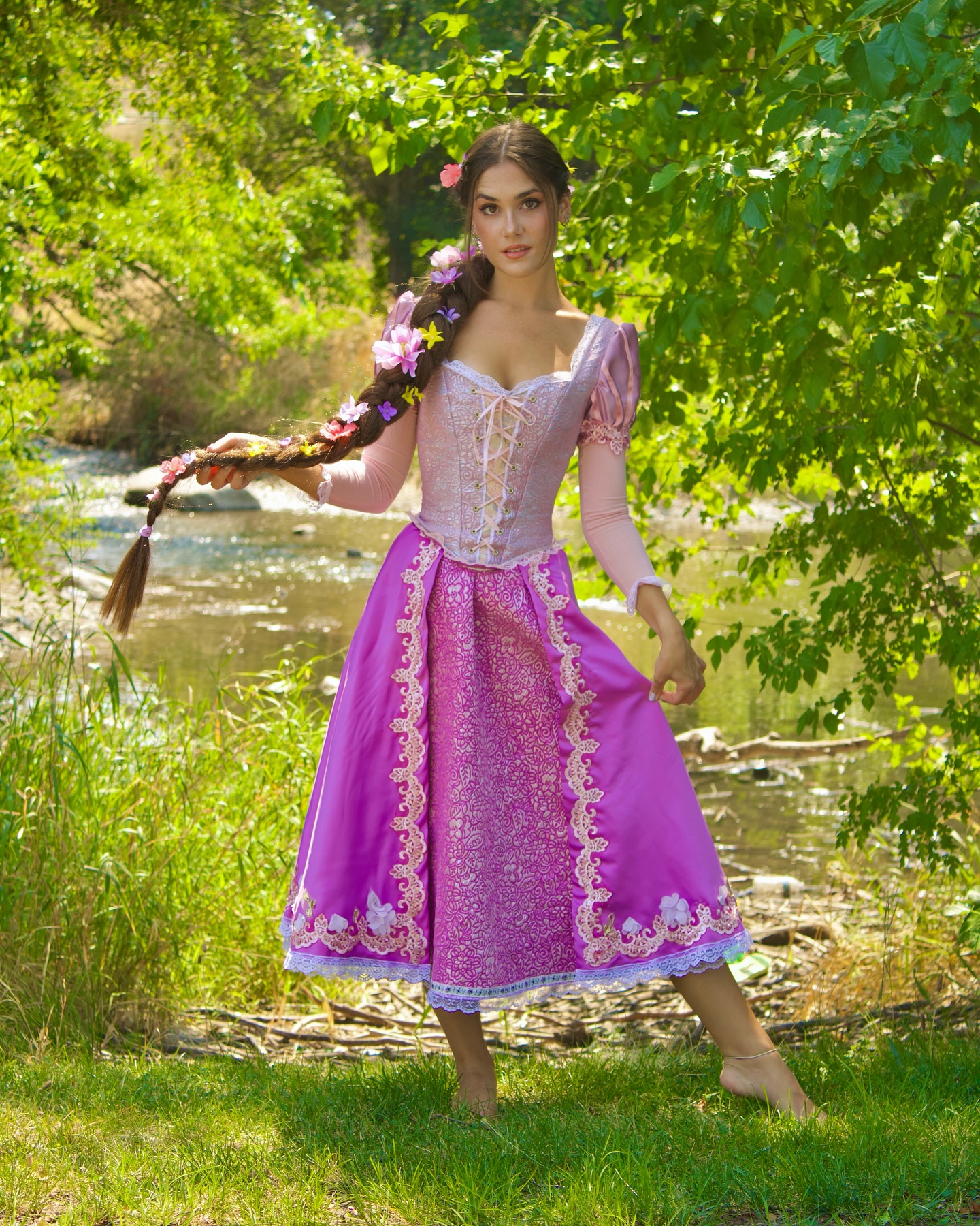 a young woman is cosplaying as rapunzel from disney's tangled. she is wearing a beautiful purple dress with a corset top and a long, flowing skirt. her long brown hair is braided and adorned with flowers. she is standing in a natural setting by a river, barefoot, and looking directly at the camera. the overall image is bright and whimsical, capturing the essence of a fairytale princess.
