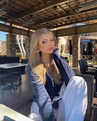 the image shows loren gray sitting in an outdoor restaurant patio. she has blonde hair and is wearing a blue jacket and white pants. the patio has stone columns and wicker chairs. there are tables and chairs set up for outdoor dining.
