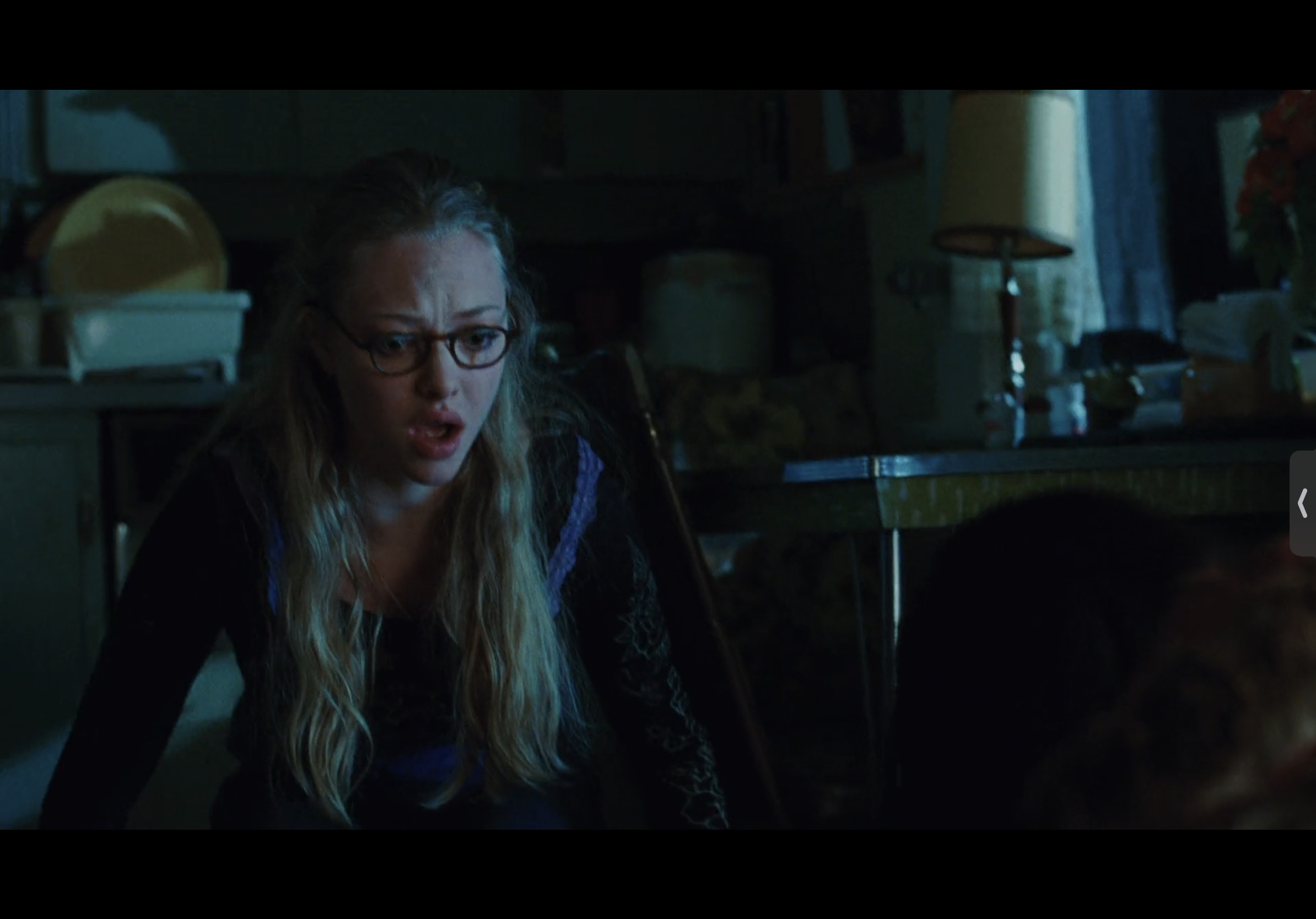a young blonde woman wearing glasses looks scared while sitting in a dimly lit room. she is dressed in a black and purple top and her long blonde hair is over her shoulders. she is looking toward the camera. the room appears to be a kitchen, with a table, lamp, and other furniture in the background. the image is dark and the woman's facial expression suggests she is afraid or worried.