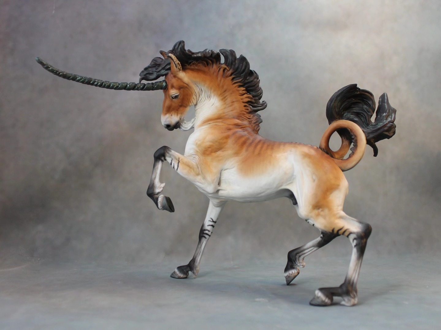 a detailed sculpture of a brown and white unicorn with a black mane and a curly tail, standing on a gray surface. the unicorn has a long, textured horn and black markings on its legs.