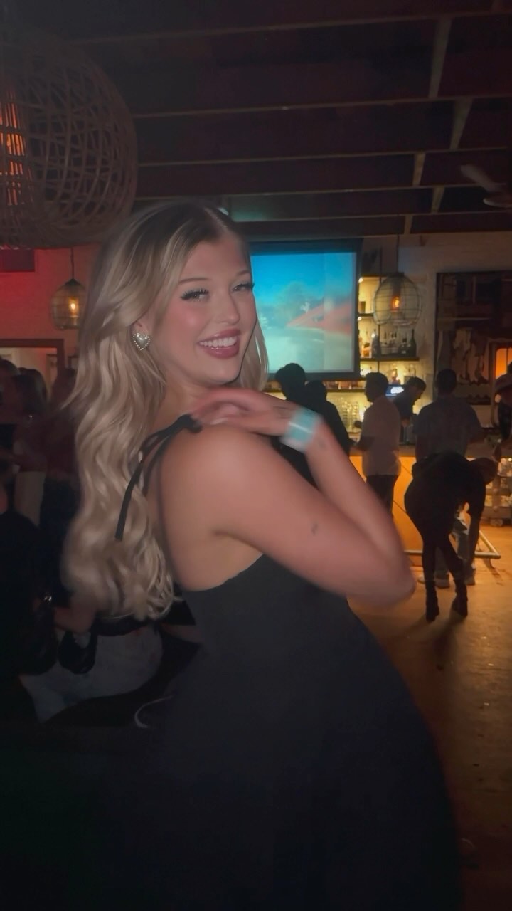 a young woman with blonde wavy hair and a black dress is smiling at the camera. she is indoors, possibly in a bar. there are other people in the background.