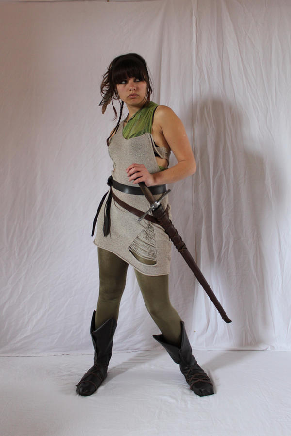 a fullbody studio shot of a young woman dressed in a medievalinspired warrior costume. she wears a ripped beige tunic over a green undershirt, olive green tights, and dark brown boots.  she is holding a sword at her hip, and her gaze is directed away from the camera. the background is a simple white backdrop.  her hair is styled loosely and she has a small feather tucked into it. the overall aesthetic suggests a fantasy or roleplaying character.