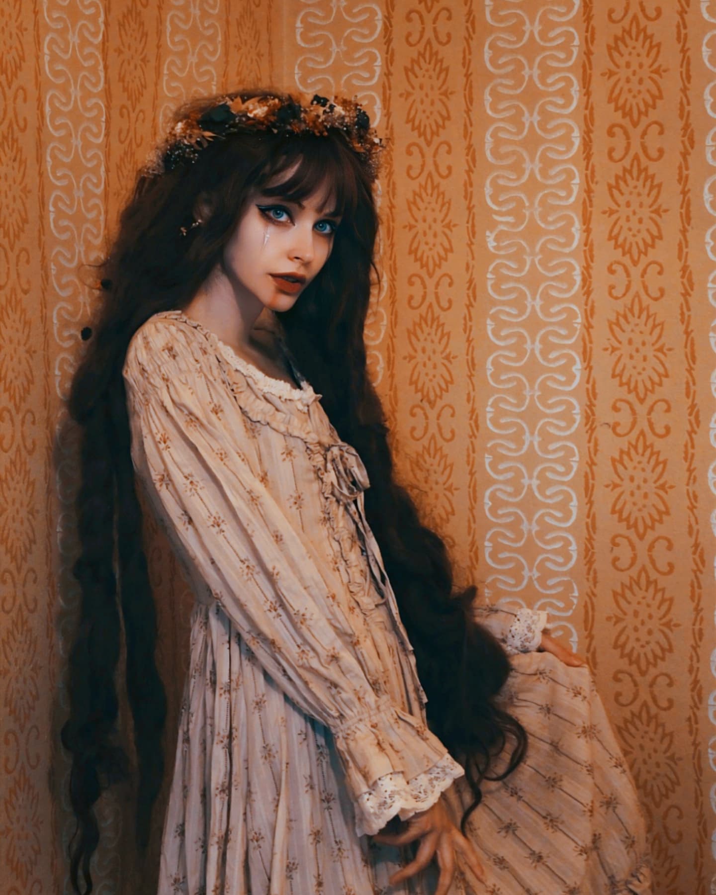 a young woman with long dark hair adorned with a floral crown is captured in a portrait. she is wearing a vintagestyle, light beige floral dress with lace detailing. the dress has long sleeves and a slightly fitted bodice. the woman's makeup emphasizes her dark eyes and pale skin, creating a somewhat ethereal look. she is standing against a backdrop of an orange vintagestyle wallpaper with a repeating white floral pattern. the overall mood is romantic, mystical, and slightly gothic. the lighting and composition of the image contribute to a dreamy, artistic feel.