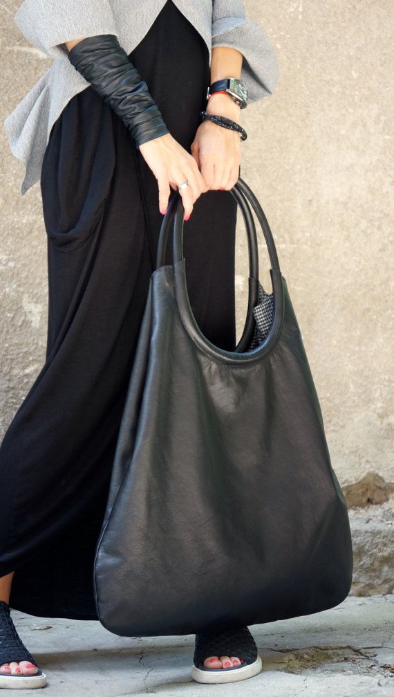 a woman is holding a large black leather bag, dressed in a long black dress with a gray jacket and leather sleeves. she is wearing black sandals and accessorized with a watch, bracelet, and ring. the background is a neutral wall.