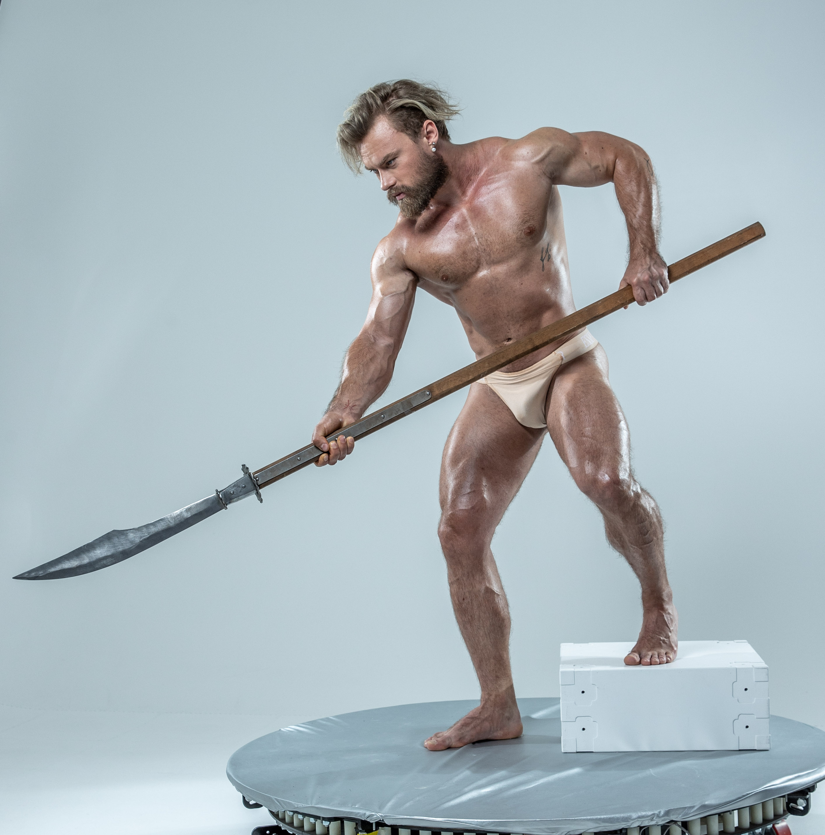 a muscular man with blond hair in a warriorlike pose holds a spear. he's standing on a round platform against a light gray backdrop in what appears to be a studio setting. the image is professionally lit and taken from a low angle, which emphasizes the man's physique and the power of his pose.