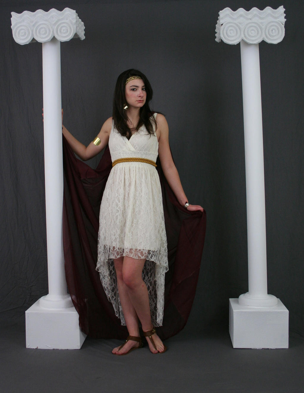 a young woman in a white lace highlow dress and maroon cape stands between two white classical columns against a gray background. she is wearing gold jewelry and sandals, and her hair is styled simply. the image is a fashion photograph, possibly intended to evoke a classical or historical aesthetic.