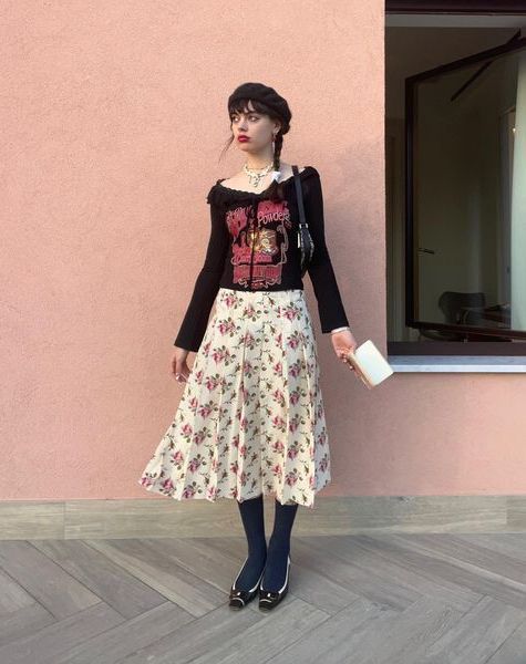 a fullbody shot of a young woman posing outdoors in front of a pink wall. she is wearing a black offshoulder cardigan over a band tee, a pleated floral midi skirt, dark tights, and dark flats. she is carrying a small white handbag and accessorized with a necklace. the overall style is a mix of vintage and modern elements.