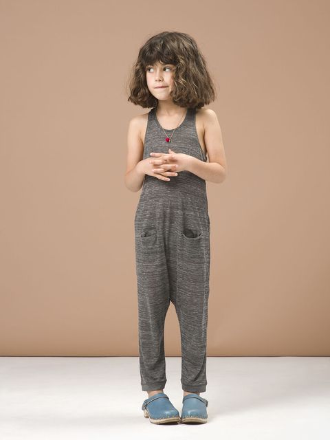 the image shows a young girl with shoulderlength curly brown hair wearing a heather grey jumpsuit. the jumpsuit has a sleeveless tank top style and loosefitting pants with small pockets. she is also wearing light blue clogs and a small red pendant necklace. the girl is standing against a light beige backdrop and appears to be in a studio setting.  the overall style is casual and comfortable. the image appears to be from a children's clothing catalog or similar promotional material.