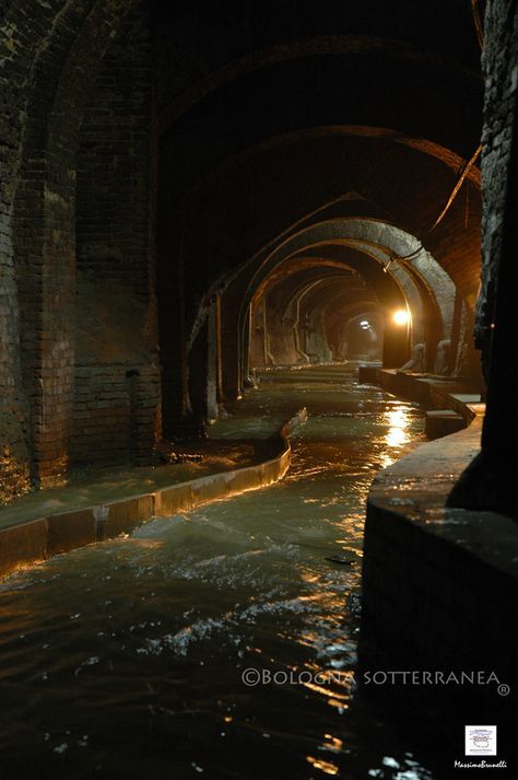 this image shows a dimly lit underground canal with arched brick tunnels. the canal is filled with water, and the brick walls create a series of repeating arches leading into the distance. the scene is dark and mysterious, with shadows obscuring some of the details. the image is labeled "bologna sotterranea," suggesting that this is a subterranean canal system in bologna.