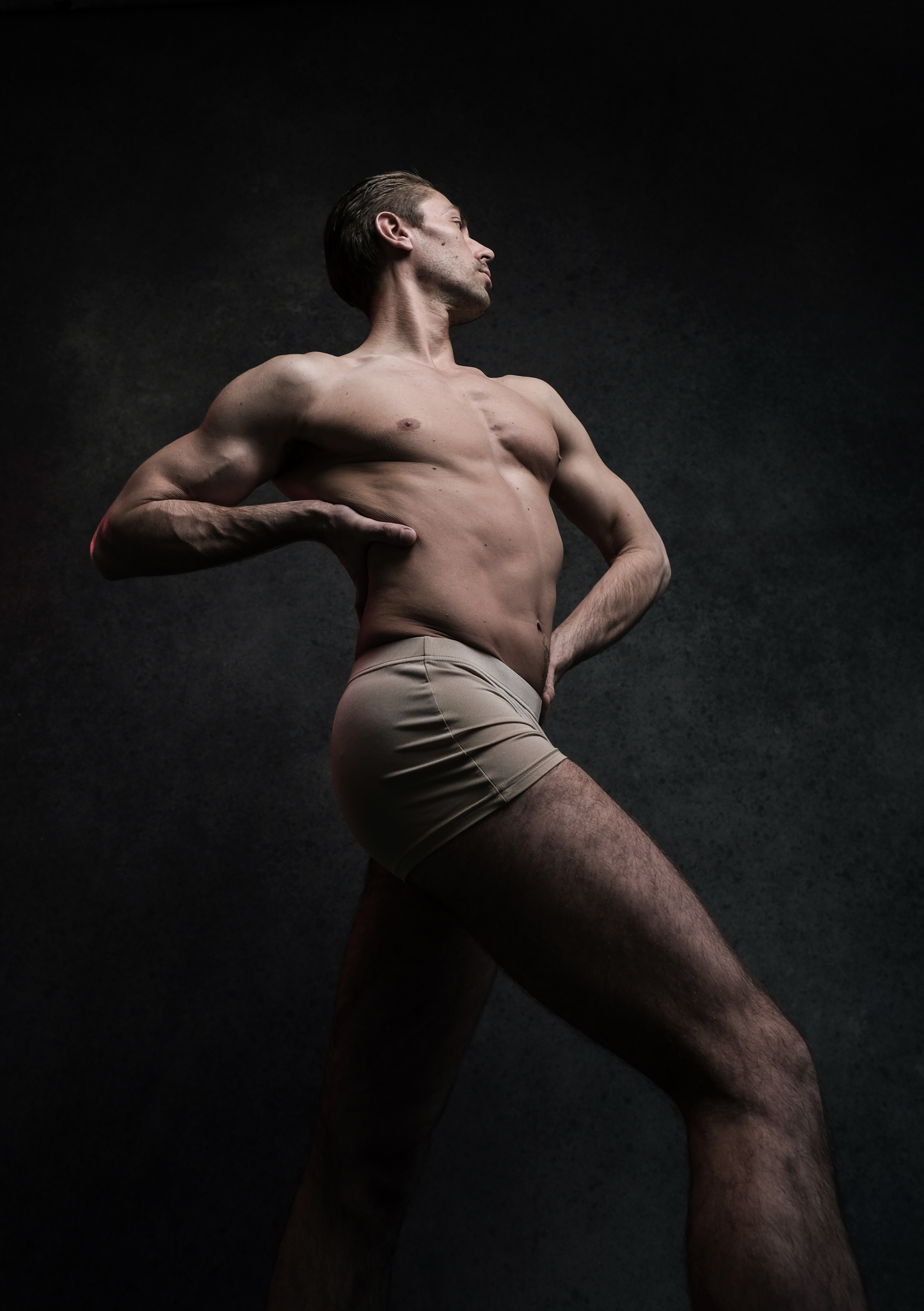 a muscular man in beige underwear stands against a dark gray background. the studio lighting casts dramatic highlights and shadows on his body, emphasizing his physique. he stands in a graceful pose, with his head tilted back and his left hand resting on his hip. the lowangle perspective adds to the sense of power and strength.