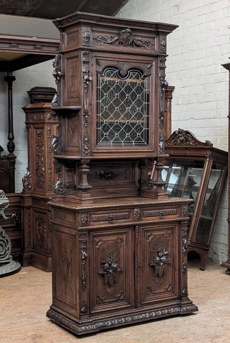 a detailed photograph features an antique, elaborately carved wooden cabinet. the cabinet is made of dark wood, likely oak or mahogany, and stands tall with multiple sections. the bottom part of the cabinet has doors with intricately carved floral designs. above this is a countertop area, and then another section with a glassfronted display case. the glass is divided into a diamond pattern, adding to the antique aesthetic. the cabinet is heavily ornamented with carvings, including floral patterns and possibly animal figures. it appears to be a combination of a buffet and a hutch. the surrounding area shows other pieces of furniture, possibly in a showroom or antique shop. the lighting is slightly subdued, adding to the vintage feel of the piece.