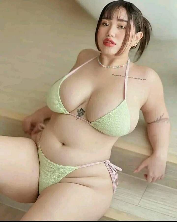 a woman with dark hair is posing while sitting on stairs wearing a light green bikini. she has tattoos on her chest and arm, and is wearing jewelry around her neck and earrings.