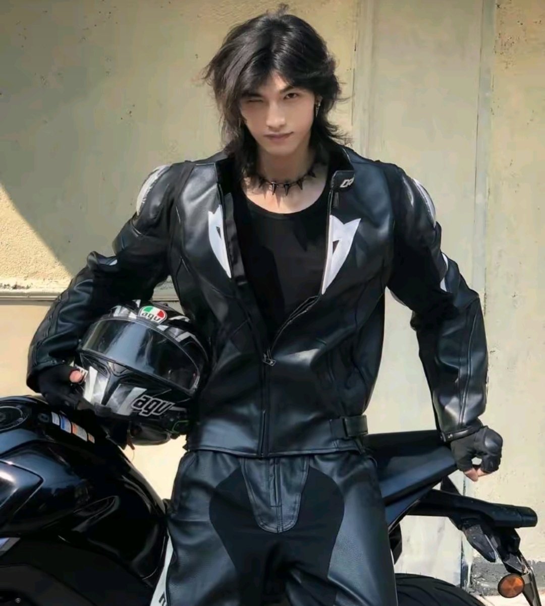 a young man wearing a black leather jacket and matching pants stands with a black motorcycle. he has a helmet in his hand, his hair is long and black, and he is wearing a black necklace. he appears to be in his early twenties and is very handsome and stylish.