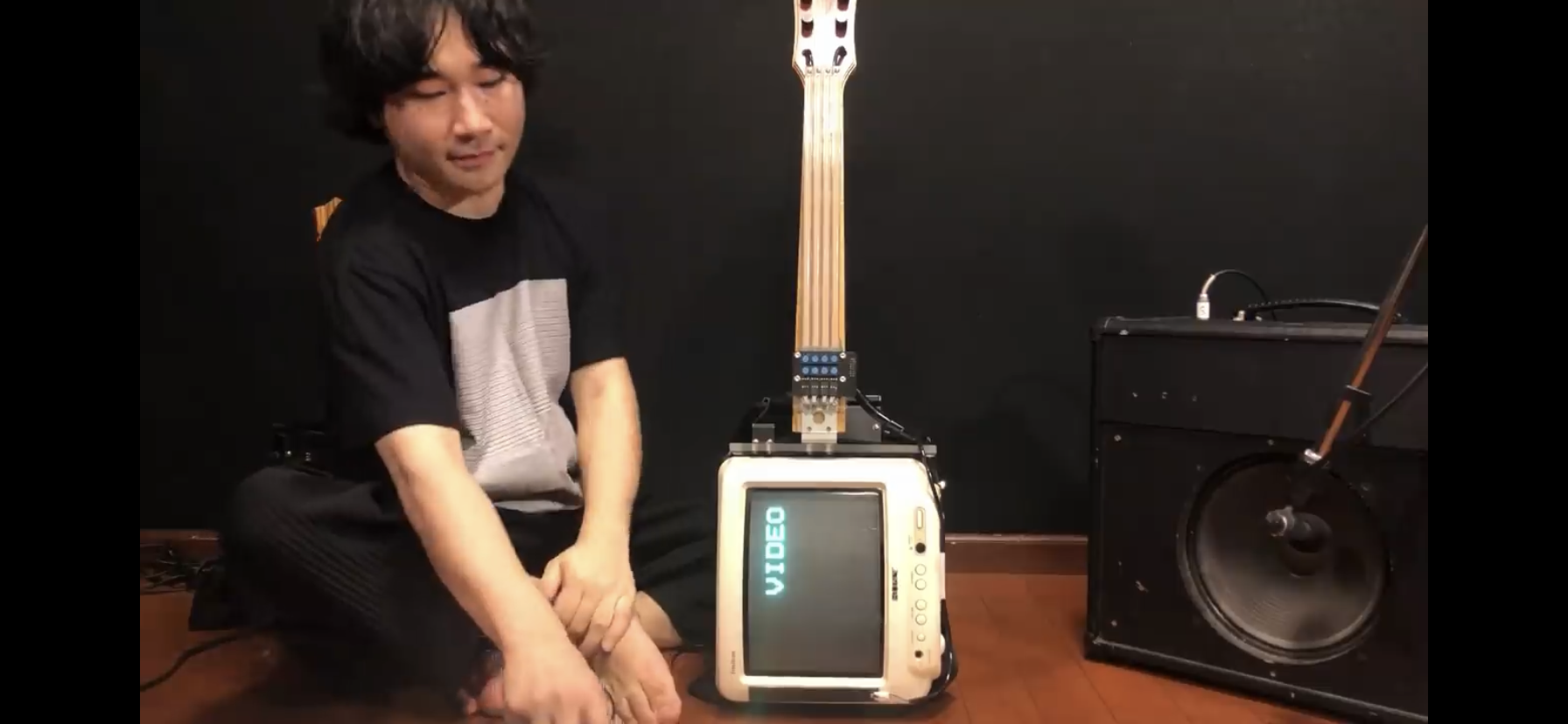 a man is sitting on the floor in front of a unique bass guitar with a small crt television as its body. the television screen displays the word "video" in blue letters. the guitar is plugged into a small amplifier to the right. the man is looking at the camera with a neutral expression. he is wearing a gray and white shirt and white pants. the room is dimly lit with warm artificial light.  the walls are dark, and the floor appears to be hardwood.