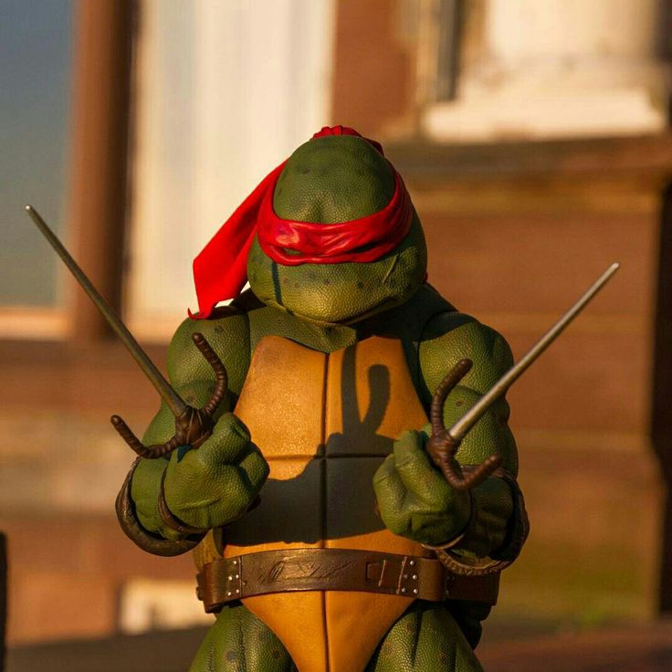 a teenage mutant ninja turtle action figure stands ready for battle, wielding a pair of sais. the figure is dressed in its signature green and orange garb, with a red bandana over its eyes. it is standing in front of a brown and yellow backdrop, ready to strike.