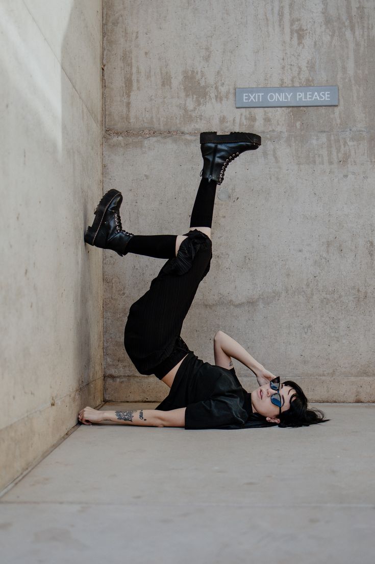 the image showcases a young woman lying upside down in a corner formed by a grey concrete wall.  she is dressed in all black attire; a black tshirt, black pants and black combat boots. one leg is bent and the other is extended straight in the air, almost touching the wall. she wears sunglasses and has visible tattoos on her arm. the overall style is edgy and urban, with a cool, rebellious attitude. a small "exit only please" sign is visible on the wall. the scene is minimalist and gritty, enhancing the fashion and pose of the model.