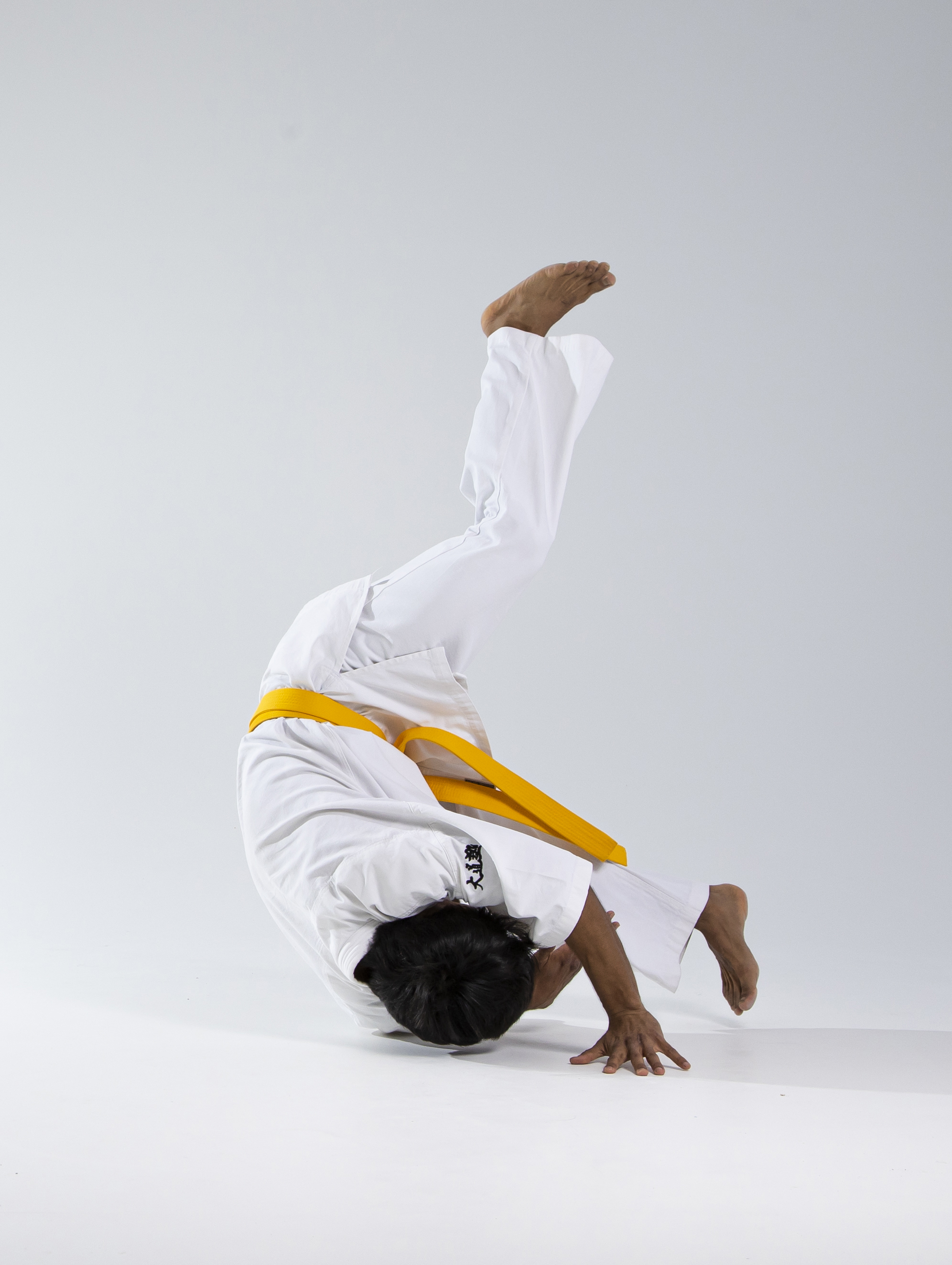a person in a white karate uniform with a yellow belt is performing a headstand. the individual's head is on the ground, supporting their weight, and their legs are extended upwards. they are in a white studio setting.