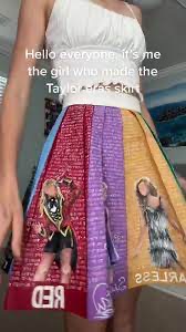 a woman with a white tank top is standing with her right hand on her hip. she is wearing a multicolored skirt with different taylor swift eras on it. the skirt has red, purple, yellow, and blue stripes.  the skirt is made of fabric with printed text on each color panel. the woman is smiling at the camera.