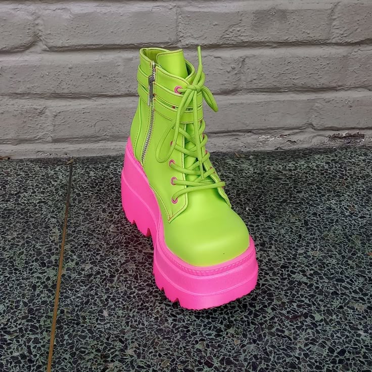 a single neon green platform boot with bright pink sole is displayed on a speckled grey surface. the boot has laces and a zipper. the background is a light grey brick wall.