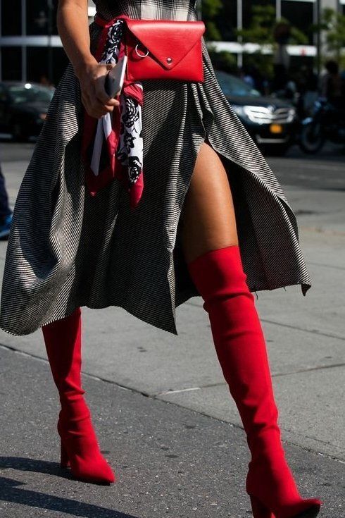 a woman is walking down a city street, showcasing her stylish outfit. she's wearing a black and white plaid skirt with a high leg slit, a red belt bag, and a red silk scarf. her footwear is a pair of striking red overtheknee boots. the overall look is chic, trendy, and sophisticated. the background is slightly blurred but shows other people and city elements. the image captures a moment of street style fashion.