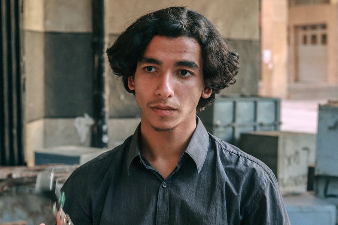 a closeup portrait of a young man with dark, curly hair. he is wearing a dark gray buttondown shirt and has a serious, somewhat pensive expression on his face. the background is blurred but shows an urban setting with concrete structures and storage containers. the image has a somewhat moody and atmospheric quality. the young man appears to be of middle eastern or arabic descent.
