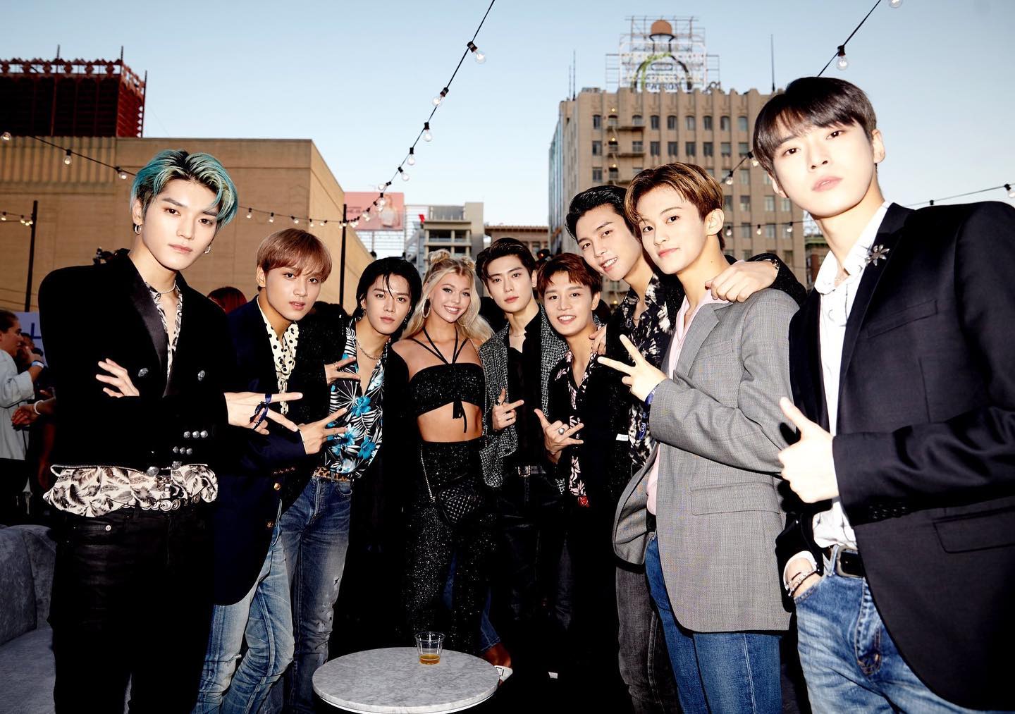 a group of people, including a blonde woman and members of a korean pop group, are posing for a photo on a rooftop with cityscape and buildings in the background. string lights are strung overhead. the group members are dressed in fashionable outfits, including suits and patterned shirts. they are making hand gestures.