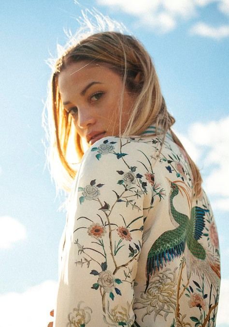 a young woman with blonde hair is captured outdoors against a backdrop of blue sky and fluffy white clouds. she wears a white satin bomber jacket adorned with intricate floral and bird embroidery, showcasing a blend of elegance and natureinspired style. the lighting is bright and sunny, highlighting the details of her jacket and creating a warm, summery atmosphere. the perspective is over her shoulder as she glances back at the camera, adding a sense of intrigue to the portrait.
