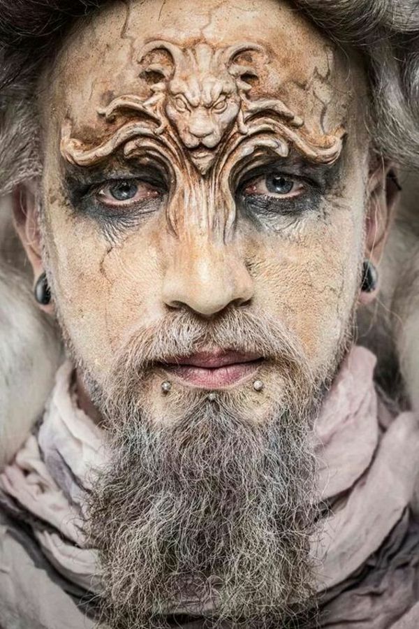a closeup portrait of a man with elaborate fantasy makeup. a sculptural lion design is applied to his forehead. he has gray hair, a gray beard, and lip piercings. his eyes are accented with dark makeup. he is wearing a white fur shawl and a draped lightcolored shawl. the overall style is dramatic and otherworldly.
