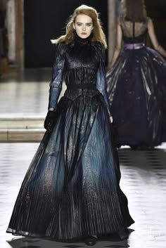 a fashion model walks down a runway in a black and silver long gown. the dress is very elegant and is accented by a blue hue. the model is wearing the dress beautifully and has a confident pose. the backdrop of the runway is a blurred image of the audience and a white wall.