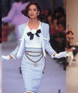 the image showcases a runway model from the 1990s, possibly a chanel show. she's wearing a classic chanelesque light blue and white tweed skirt suit with a white fitted top underneath. a large black bow adorns the neckline, and she's accessorized with multiple strands of a pearl and gold chain necklace, and white gloves. her hair is styled in a short, slightly tousled bob.  the background is blurry, focusing attention on the model and her attire, showcasing the high fashion of the era. the image appears to be a scan from a magazine or other print publication.