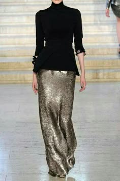 a fashionable woman wearing a long, black turtleneck sweater with a gold sequined skirt. she is walking down a runway, posing for the cameras.  the woman looks elegant, stylish, and glamorous.
