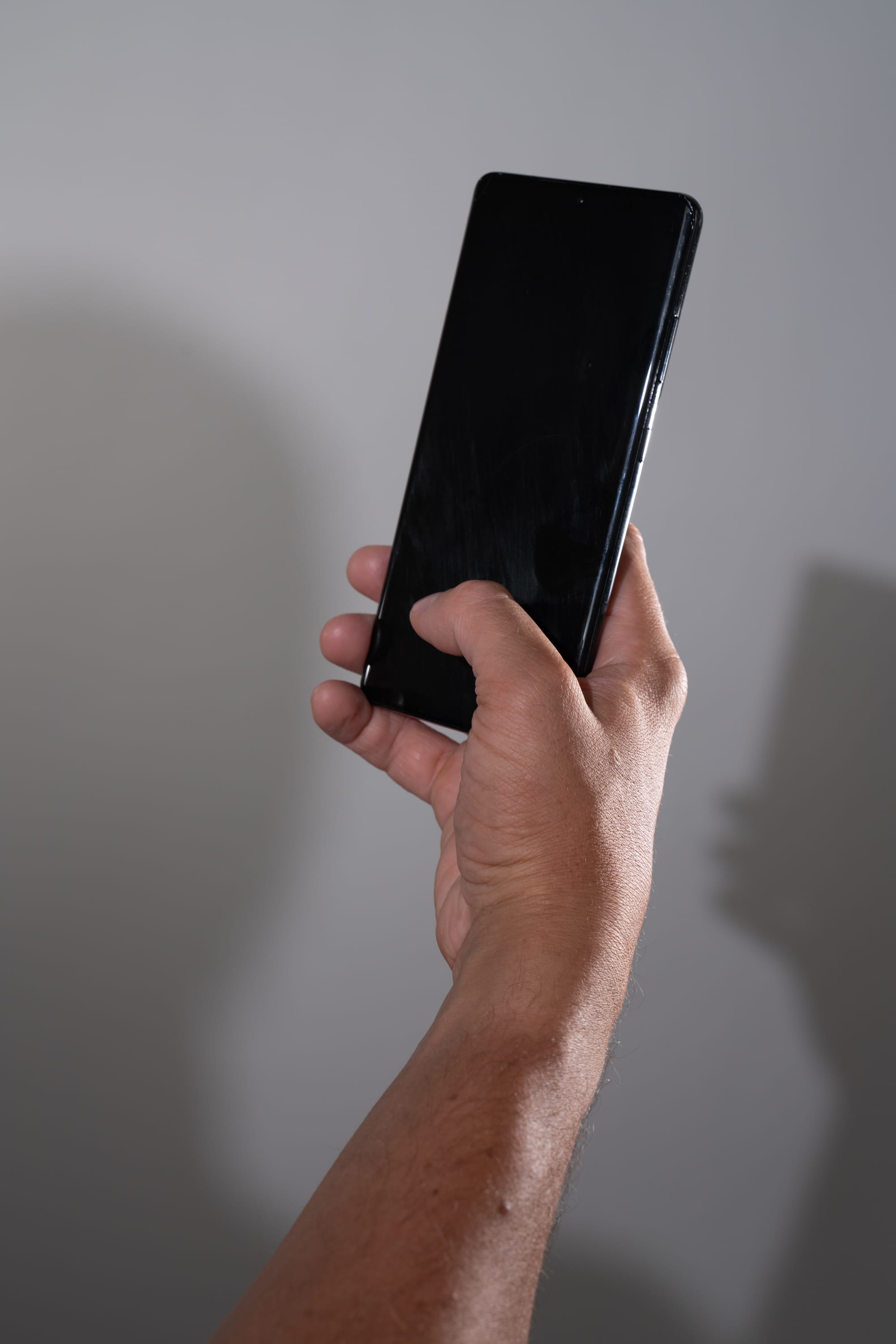 the image shows a man’s hand holding a black cell phone against a gray background. the screen of the phone is black. the photo is taken from a worm's eye view, looking up the man’s arm towards the phone. the lighting is soft and casts a shadow of the man’s hand and phone on the wall behind.