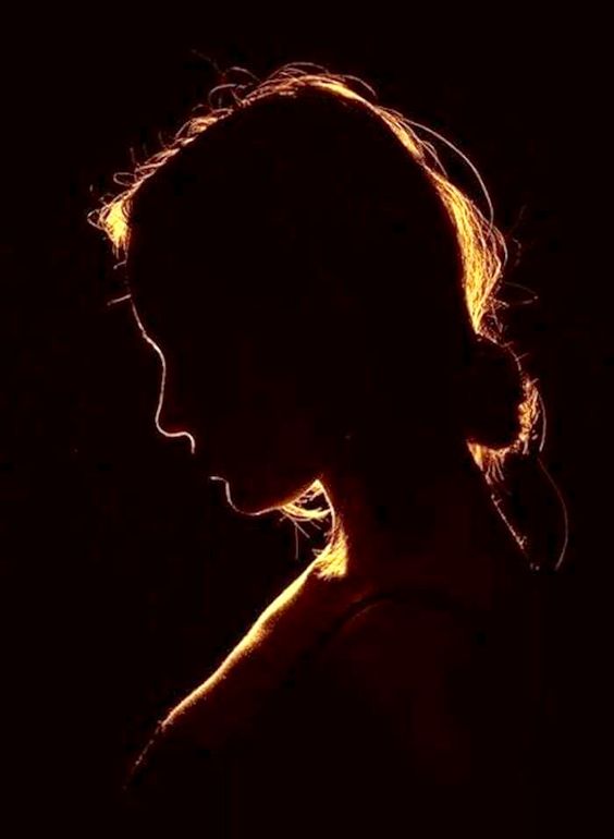 the image shows the silhouette of a woman's profile against a dark background. her hair is pulled back, and her head is slightly bowed. the lighting is dramatic, with a rim light outlining her hair and shoulders, creating a halo effect. the overall mood of the image is contemplative and mysterious.