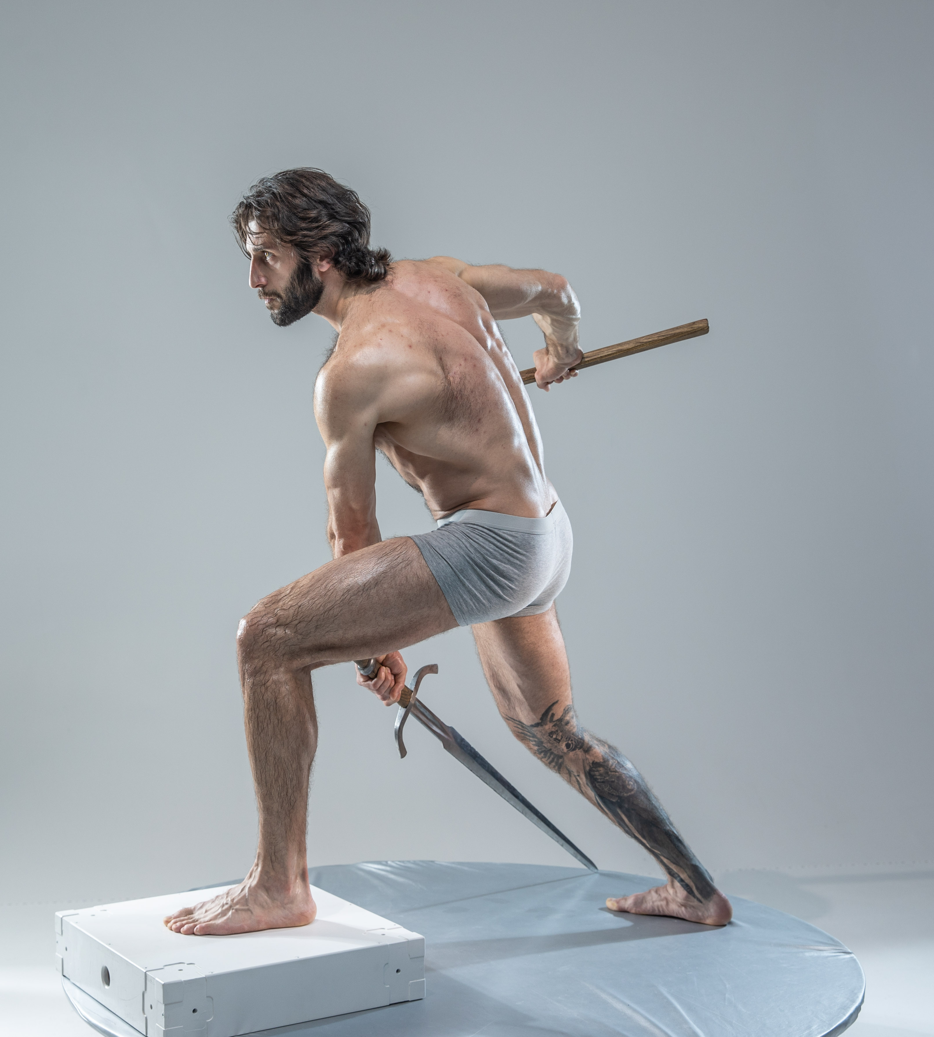 a muscular man with long hair and tattoos is posed dynamically against a light gray background in a studio setting. he is holding a sword in his right hand and a wooden staff in his left hand, with his body slightly turned. he is wearing grey underwear and his physique is welldefined, suggesting an athletic build. the lighting is soft and diffused, highlighting the contours of his body. the overall image evokes a sense of action and strength.