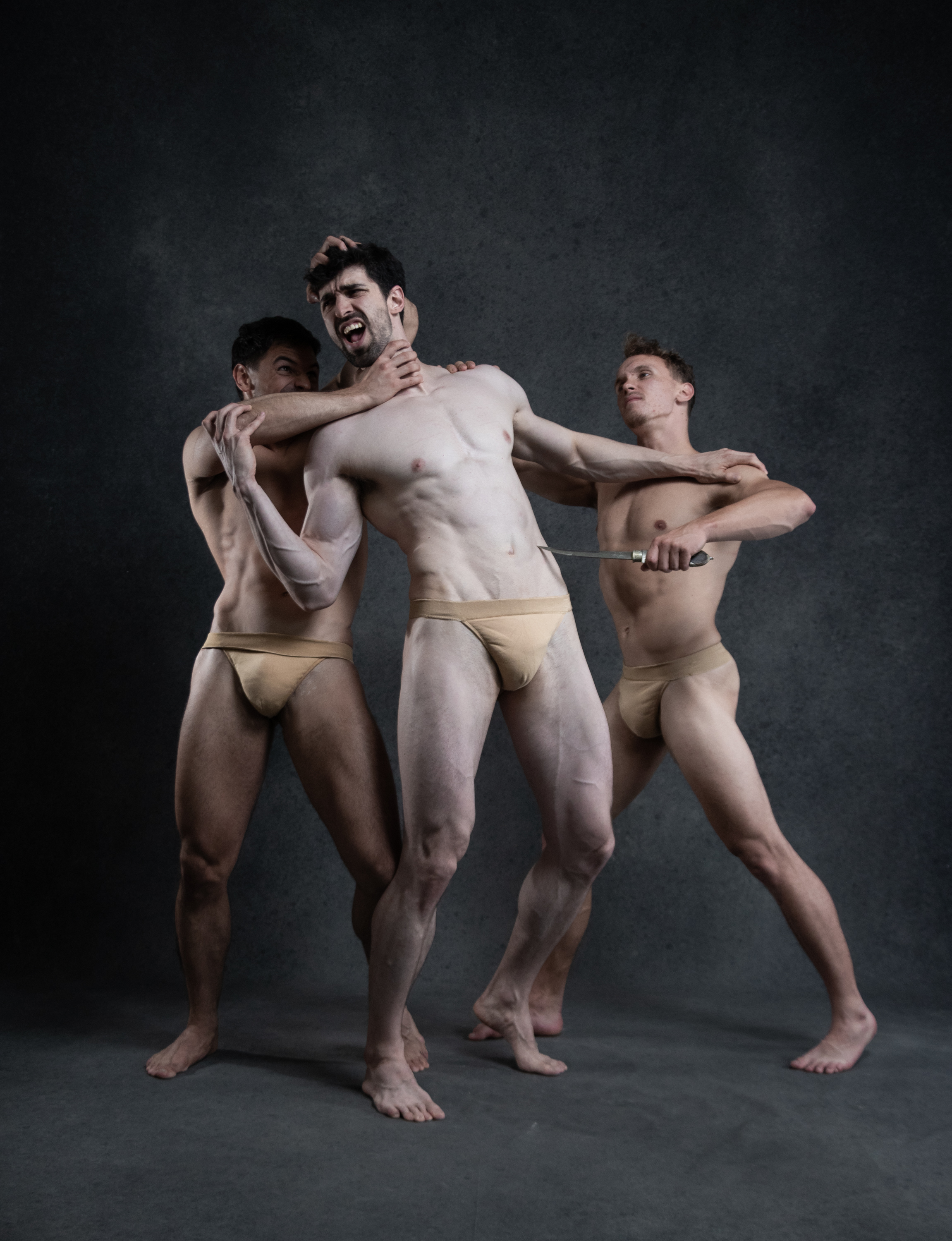 three muscular men are locked in a fierce struggle against a dark gray backdrop. two of the men have grabbed hold of the central figure, who appears to be in distress. one man has his hand around the central man's throat, while the other brandishes a knife menacingly. the image evokes a sense of danger, aggression, and raw physicality.